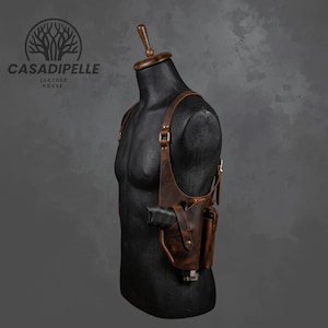 May include: A brown leather shoulder holster system featuring adjustable straps, metal hardware, and a moulded pouch for a firearm. The holster is displayed on a black mannequin torso with the CASADIPELLE LEATHER HOUSE logo visible in the upper left corner.