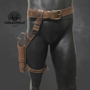 May include: A brown leather thigh holster belt with a waist strap, a vertical leg strap, and a cylindrical pouch. The belt features a metal buckle and the text CASADIPELLE LEATHER HOUSE with a tree logo printed on the upper left side of the image.