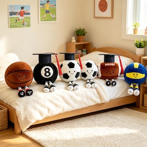 May include: A collection of six plush sports balls including a basketball, an eight ball, two footballs, an American football, and a volleyball. Each plush toy features a smiling face, dangling legs with numbered feet, and a black graduation cap with a red tassel.
