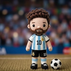 Puede incluir: Una muñeca de ganchillo que representa una figura de pelo castaño con barba, que lleva una camiseta a rayas blancas y azul claro con el número 10 y un emblema, pantalones cortos negros con el número 10 y un pequeño balón de fútbol.