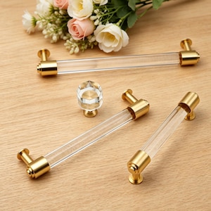 Pode incluir: Três puxadores de armário em acrílico transparente com extremidades em metal dourado escovado e um puxador hexagonal a condizer em acrílico transparente. As ferragens apresentam um design de barra cilíndrica com suportes dourados.