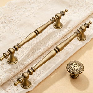 Puede incluir: Dos tiradores de cajón de metal en tono latón con un eje central liso y cónico y detalles decorativos de anillos en los extremos, acompañados de un pomo de armario redondo con un patrón floral radiante y detallado en su cara.