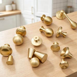 May include: A collection of brass-toned cabinet hardware including various knobs, pulls, and handles. The set features smooth spherical, ribbed, and conical shapes with a matte metallic finish, designed for furniture or cabinetry installation.