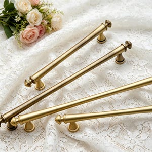 May include: Four long, cylindrical cabinet pulls with a brushed brass finish. Each handle features a smooth, straight bar design with decorative, rounded finials at both ends and two small, conical mounting posts for attachment to furniture.