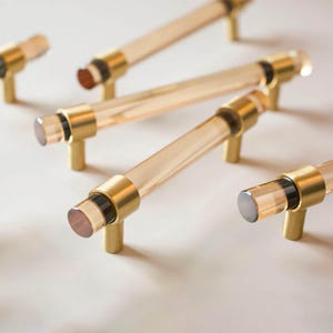 May include: A collection of cabinet pulls featuring clear, cylindrical acrylic bars accented with brushed gold metal mounts and black circular spacers at each end. The hardware components are arranged in a group, showcasing the combination of transparent material and metallic finish in a functional design.