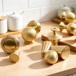 May include: A collection of gold-toned metal cabinet knobs and pulls in various shapes, including spherical, ribbed, and conical designs. These hardware pieces feature a smooth, brushed finish and are arranged in a group on a light wooden surface.