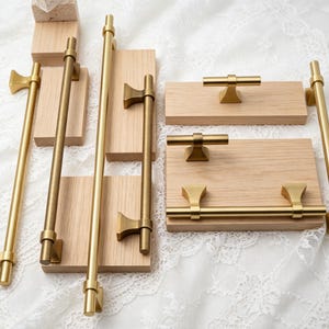 Puede incluir: Una colección de herrajes para armarios de metal dorado cepillado, que incluye tiradores de barra largos y pomos en forma de T, montados sobre bloques de madera rectangulares de color claro. Los herrajes presentan un diseño cilíndrico liso con bases trapezoidales ensanchadas.