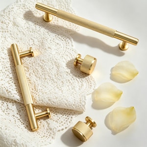 May include: Two gold-toned metal cabinet pulls and two matching round cabinet knobs, each featuring a textured, ribbed center section. The hardware pieces are arranged with a white lace fabric and three pale yellow flower petals.