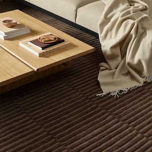 May include: A dark brown area rug with a textured, ribbed, linear pattern. The rug is positioned beneath a light-colored sofa, a light wood coffee table, and a beige throw blanket with fringe, creating a neutral-toned living space.