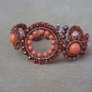 May include: A handcrafted bracelet featuring copper-colored wire woven into circular and floral designs. The bracelet incorporates peach-colored beads and accents, with a silver-toned clasp. The intricate design showcases a unique, artistic style.