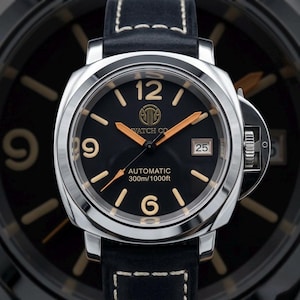 May include: A stainless steel wristwatch with a black face, featuring gold-coloured hands and numerals. The watch has a date display at the 3 o'clock position and is mounted on a black leather strap with white stitching. The watch face reads "WATCH CO" and "AUTOMATIC 300m/1000ft".