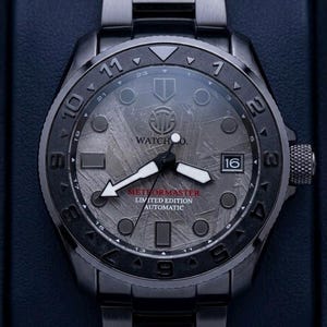 May include: A dark gray wristwatch with a textured face and a matching metal band. The watch face features the text "WATCH CO." and "METEORMASTER LIMITED EDITION AUTOMATIC". The date is displayed at the 3 o'clock position.