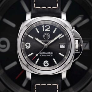 Puede incluir: Un reloj de pulsera de acero inoxidable con esfera negra y correa de cuero negro. La esfera presenta números blancos, una ventana de fecha y el texto "AUTOMATIC 300m/1000ft". La correa está cosida con hilo blanco.