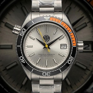 May include: A stainless steel wristwatch with a silver face, black and orange bezel, and a date window at the 3 o'clock position. The watch features a silver band and a yellow second hand. The watch face has the text "ATTA" and "SUPERLATIVE CHRONOMETER OFFICIALLY CERTIFIED."