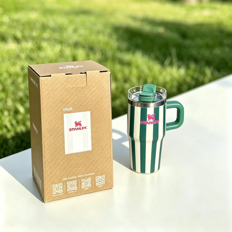 May include: A green and white striped Stanley travel mug with a green handle and lid, next to a cardboard box with the Stanley logo. The mug has a pink logo and is on a white surface with a green background.