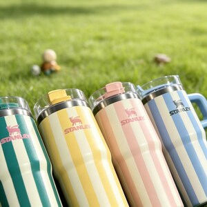 May include: Four Stanley insulated tumblers with vertical stripes in green, yellow, pink, and blue. Each tumbler has a clear lid and a matching colored handle or top. The Stanley logo is visible on each tumbler.