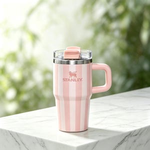 May include: Pink and white striped Stanley travel mug with a handle and a clear lid. The mug has the Stanley logo and a pink flip-top closure. The mug is on a white marble surface.