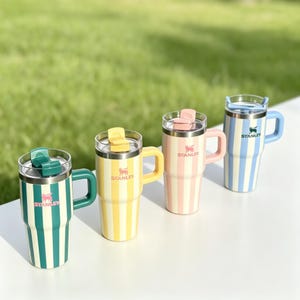 May include: Four insulated travel mugs with vertical stripes in green, yellow, pink, and blue. Each mug has a matching handle and lid, with the brand name "Stanley" and a logo. The mugs are on a white surface with a green background.