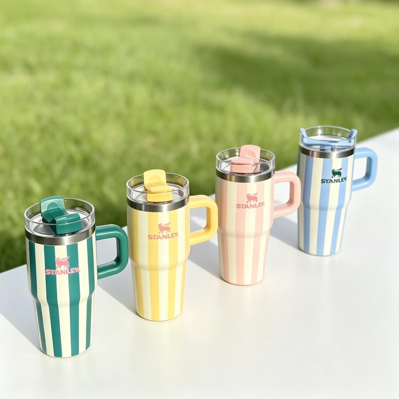 May include: Four insulated travel mugs with vertical stripes in green, yellow, pink, and blue. Each mug has a matching handle and lid, with the brand name "Stanley" and a logo. The mugs are on a white surface with a green background.