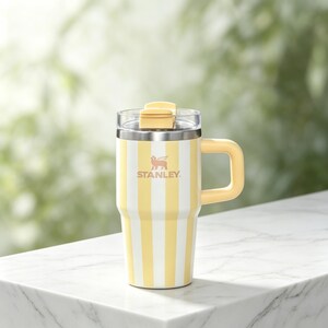 May include: Yellow and white striped Stanley travel mug with a handle and a clear lid. The mug has the Stanley logo in the center. The handle and the top of the lid are yellow.