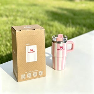 May include: A pink and white striped Stanley travel mug with a handle and a lid. The mug is next to a cardboard box with the Stanley logo. The mug has a pink handle and a pink lid. The box has the Stanley logo and the word "COLOR" printed on it.
