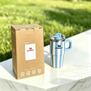 May include: A blue and white striped Stanley travel mug with a handle and lid, next to its cardboard box. The box features the Stanley logo and product information. The mug is on a white surface with a green background.