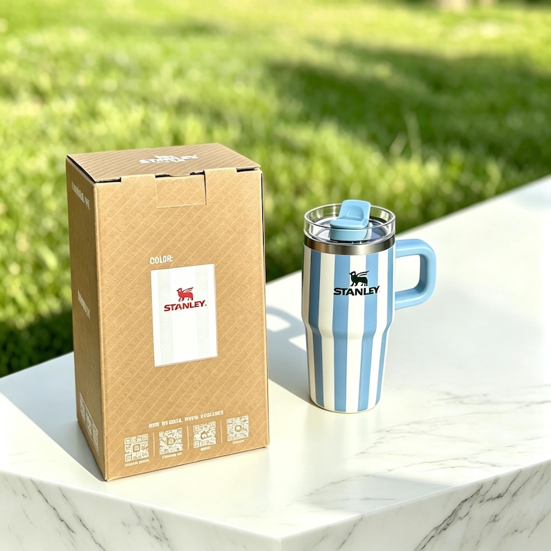 May include: A blue and white striped Stanley travel mug with a handle and lid, next to its cardboard box. The box features the Stanley logo and product information. The mug is on a white surface with a green background.