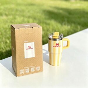 May include: Yellow and white striped Stanley travel mug with a handle and clear lid. The mug is next to its cardboard box, which has the Stanley logo and text. The mug is designed to keep beverages hot or cold.