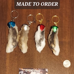 May include: Four keychains featuring gray and white fur feet with metallic bells in blue, silver, red, and green. Text reads HOLIDAY GRAY FEET MADE TO ORDER. A US quarter coin is visible for scale next to a business card with the text Double H Farms.