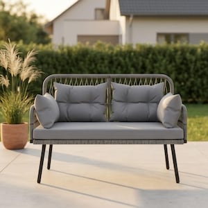 Puede incluir: Un sofá gris para exteriores con dos cojines de asiento y dos cojines de respaldo. El sofá tiene una estructura tejida y patas negras. Los cojines son de color gris liso. El sofá está sobre un patio de hormigón con un fondo de hierba.