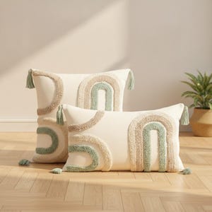 Peut inclure: Deux coussins blanc cassé avec des motifs d'arches touffetés et texturés en vert sauge et beige. Les coussins carrés et rectangulaires sont ornés de petites houppes assorties aux coins, présentant un design en relief doux sur le devant.