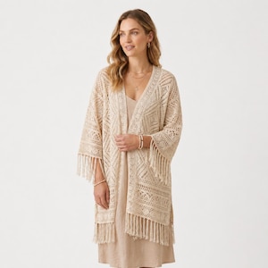 Könnte beinhalten: Eine beigefarbene, vorne offene Strickjacke mit einem komplizierten Häkelmuster aus geometrischen Rautenmotiven und langen Fransen am Saum und an den Ärmeln. Das Kleidungsstück wird über einem schlichten, einfarbigen beigen ärmellosen Kleid getragen.