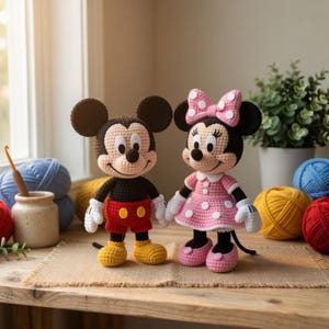 May include: Crocheted Mickey Mouse and Minnie Mouse dolls. Mickey wears red shorts, yellow shoes, and white gloves. Minnie wears a pink polka-dot dress, matching bow, and shoes. Both have black ears and smiling faces. They stand on a wooden surface.