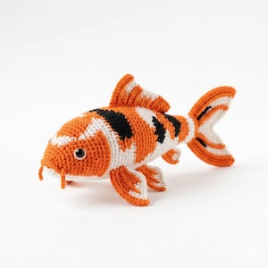 Könnte beinhalten: Ein gehäkeltes Koi-Fisch-Spielzeug mit einer strukturierten Oberfläche und einem Muster aus orangefarbenen, weißen und schwarzen Flecken. Der Fisch hat eine markante Rückenflosse, Seitenflossen, eine gegabelte Schwanzflosse und zwei kleine Barteln.