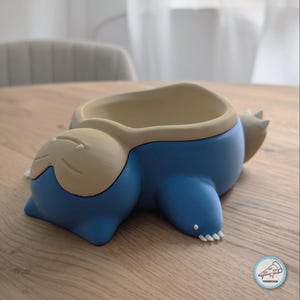 Könnte beinhalten: Ein blau-beiger Keramik-Pflanztopf in Form einer liegenden Kreatur mit geschlossenen Augen, einem kleinen Mund und weißen Krallen an den Füßen. Das Innere ist hohl und beige, und in der Ecke befindet sich ein kleines kreisförmiges Logo mit Pizzastück.