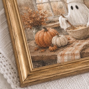 May include: A framed print featuring a ghost wearing a chef hat and stirring a bowl in a kitchen. The scene includes orange and white pumpkins, a vase of autumn leaves, and a plaid runner on a wooden table, all encased in a gold-toned frame.
