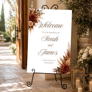 May include: A white rectangular wedding welcome sign on a black metal easel. The sign features dark red and beige floral arrangements in the top-left and bottom-right corners, with text reading Welcome to the wedding of Sarah and James, Santa Monica, CA, 08/28/2027.