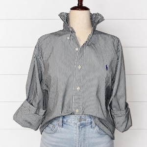May include: A long-sleeved button-down shirt featuring a black and white vertical pinstripe pattern, a high collar, and a small purple embroidered logo on the left chest. The shirt is paired with light blue denim jeans with a visible button fly.