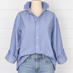May include: A light blue button-down shirt with a ruffled collar and long sleeves with folded cuffs. The shirt features a small embroidered logo on the left chest and is paired with light wash blue denim jeans, creating a casual ensemble.
