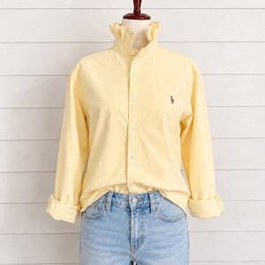 Puede incluir: Una camisa abotonada de color amarillo claro con cuello con volantes y mangas largas, combinada con vaqueros azul claro. La camisa presenta un pequeño logotipo bordado en el pecho. La letra "M" está en la esquina superior izquierda.
