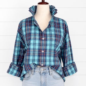 Puede incluir: Una camisa de manga larga con botones y un patrón de cuadros en tonos turquesa, azul marino y finas líneas naranjas. La camisa tiene un cuello alto con volantes, un bolsillo en el pecho y pantalones vaqueros azul claro con un botón.