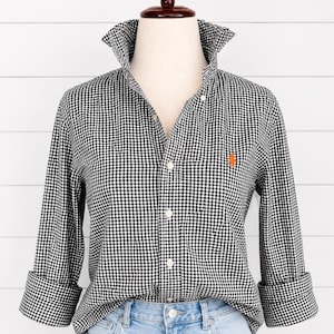 Puede incluir: Una camisa de manga larga con botones y un patrón de cuadros vichy en blanco y negro. La camisa tiene un cuello puntiagudo, una tapeta de botones frontal y un pequeño logotipo naranja bordado en el pecho izquierdo. Se combina con unos vaqueros azul claro.