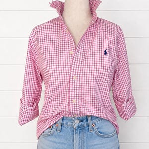 Puede incluir: Una camisa de manga larga con botones, un patrón de cuadrícula rosa y blanco, cuello alto y un pequeño logotipo azul marino bordado en el pecho izquierdo. La camisa se combina con unos vaqueros de mezclilla azul claro con botón metálico.
