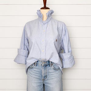 May include: A light blue button-down shirt with a ruffled high collar and long sleeves with rolled cuffs. The shirt features a small embroidered logo on the left chest and is paired with light wash blue denim jeans on a mannequin torso.