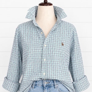 May include: A long-sleeve button-down shirt featuring a light blue, white, and dark navy blue grid pattern. The shirt has a pointed collar, a front button placket, and a small embroidered logo on the left chest. It is paired with light blue denim jeans.