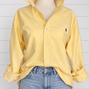 May include: A long-sleeved, light yellow button-down shirt featuring a ruffled collar and a small dark embroidered logo on the left chest. The shirt is worn tucked into light blue denim jeans, with the sleeves rolled up at the cuffs.