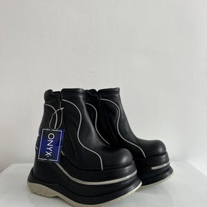 Könnte beinhalten: Ein Paar schwarze Stiefeletten mit dicker Plateausohle und hellgrauem, wellenförmigem Besatz an den Seiten. Die Stiefel verfügen über einen seitlichen Reißverschluss und ein blaues, rechteckiges Etikett mit dem weißen Schriftzug ONYX.