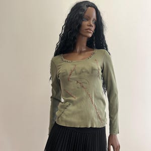 Può includere: Una maglia a maniche lunghe color verde oliva con scollo rotondo, cuciture decorative rosse visibili sul davanti e la parola "HAVE" stampata in scuro. La maglia è abbinata a una gonna plissettata nera con uno strato superiore in rete.