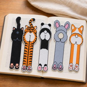 Può includere: Cinque segnalibri rettangolari a punto croce che mostrano le viste posteriori di un gatto nero, una tigre, un panda, un coniglio grigio e un corgi. Ogni segnalibro raffigura la schiena, la coda e le zampe posteriori con cuscinetti rosa.