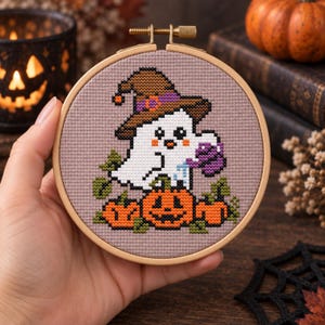 May include: A circular wooden embroidery hoop containing a cross-stitch pattern on light brown fabric. The design features a white ghost wearing a brown witch hat, holding a purple watering can, and standing behind three orange jack-o'-lanterns.